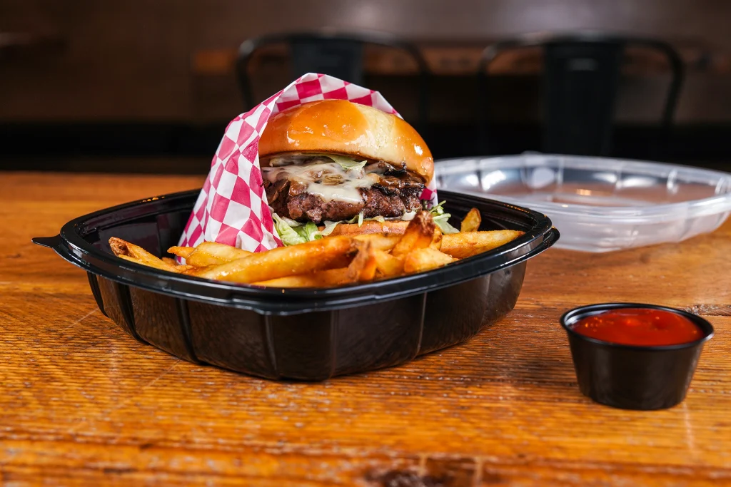 catering services, Black plastic to-go plate with mushroom and swiss burger wrapped in red and white checkered paper with a side of fries on a wood table with a side of ketchup sauce inside Farmhouse Kitchen BBQ restaurant in Sandpoint Idaho