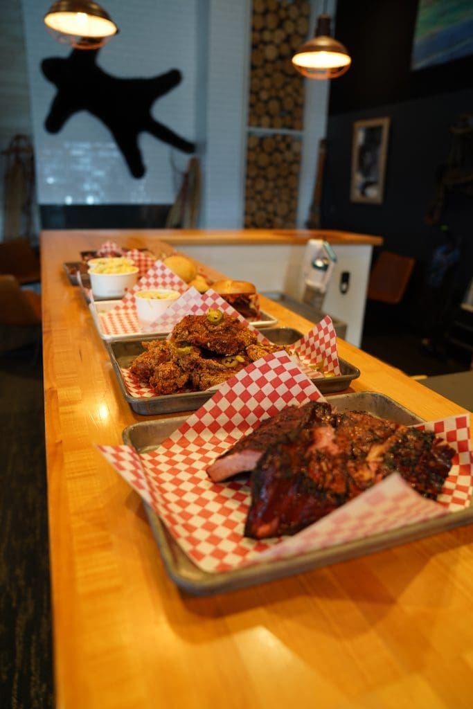 various food options on a wood counter in farmhouse kitchen bbq in sandpoint idaho with chicken and ribs on red and white checkered pattern paper in metal trays on a wood counter
